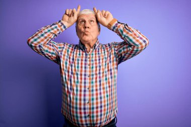 Üst düzey yakışıklı, parlak gömlekli, soyutlanmış mor arkaplan üzerine komik jestler yapan, boynuzları üzerinde parmaklarıyla gezen bir adam.