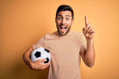 Sarı arka planda futbol topu tutan sakallı yakışıklı oyuncu şaşırmış, şaşırmış ve parmaklarıyla işaret ediyor ve kollarını kaldırıyordu..