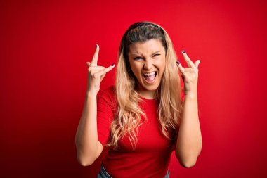Genç, güzel, sarışın bir kadın, gündelik tişört giyip izole edilmiş kırmızı arka planda dikilmiş çılgın bir ifadeyle elleri havada rock sembolü yaparak bağırıyor. Müzik yıldızı. Ağır müzik konsepti.