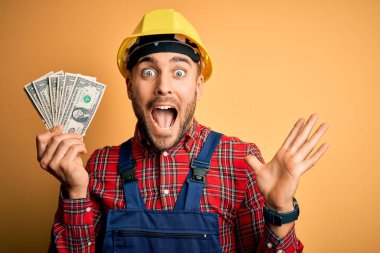 Sarı arka plan üzerinde dolar tutan kasklı genç bir müteahhit çok mutlu ve heyecanlı, kazanan yüz ifadesi zaferi büyük bir gülümsemeyle kutluyor ve ellerini kaldırıyor.