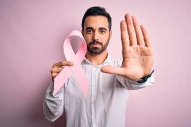 Sakallı, pembe kanser kurdelesi takan genç yakışıklı bir adam. El açık, ciddi ve kendinden emin bir ifadeyle dur işareti yapıyor. Savunma jesti.