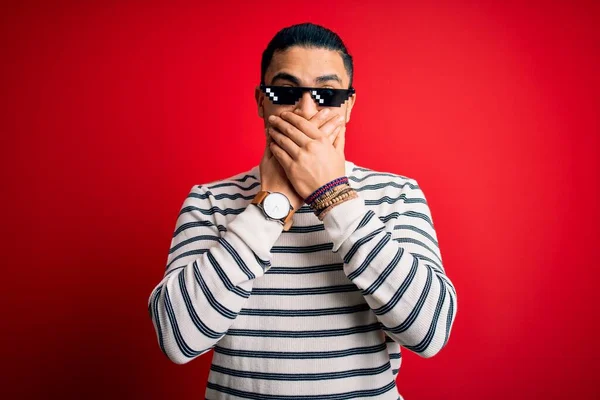 Young brazilian man wearing funny thug life sunglasses over isolated red background shocked covering mouth with hands for mistake. Secret concept.