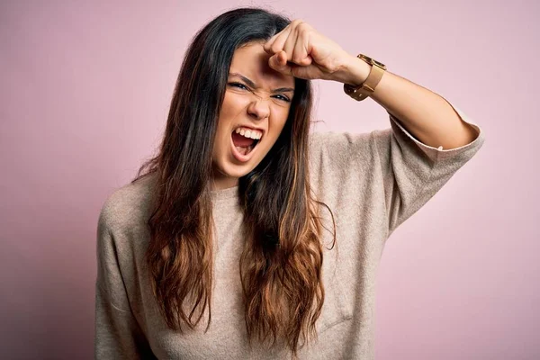 Young beautiful brunette woman wearing casual sweater standing over pink background angry and mad raising fist frustrated and furious while shouting with anger. Rage and aggressive concept.