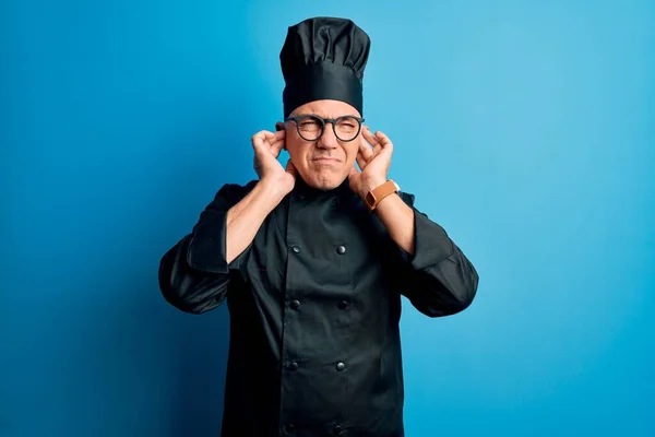 Middle age handsome grey-haired chef man wearing cooker uniform and hat covering ears with fingers with annoyed expression for the noise of loud music. Deaf concept.