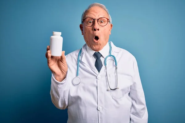 Starszy Siwowłosy Lekarz Trzymający Tabletki Farmaceutyczne Niebieskim Tle Przerażony Szoku — Zdjęcie stockowe