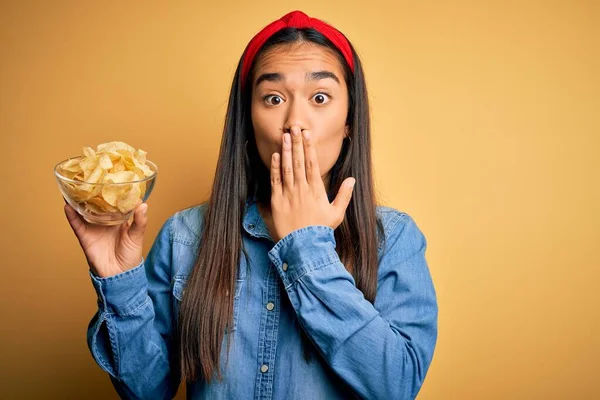 Young beautiful asian woman holding bowl with chips potatoes over isolated yellow background cover mouth with hand shocked with shame for mistake, expression of fear, scared in silence, secret concept