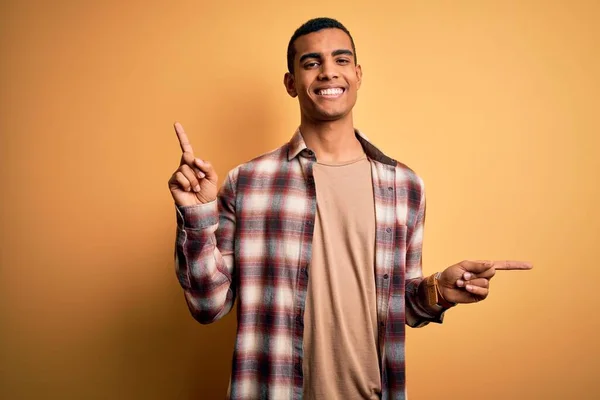 黄色の背景にカジュアルなシャツを着た若いハンサムなアフリカ系アメリカ人男性は 異なる方向に指で自信を持って指して笑顔 広告のコピースペース — ストック写真