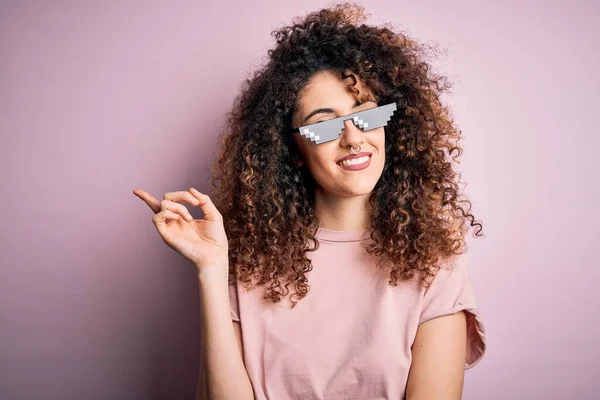 Young beautiful woman with curly hair and piercing wearing funny thug life sunglasses with a big smile on face, pointing with hand and finger to the side looking at the camera.