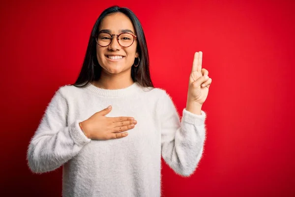 若いです美しいアジアの女性身に着けていますカジュアルセーターと眼鏡上の赤い背景笑顔身に着けています手で胸と指アップ 忠誠の誓いを作ります — ストック写真