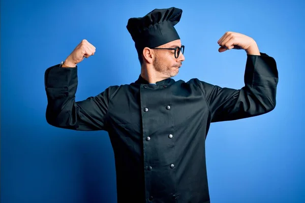 Young handsome chef man wearing cooker uniform and hat over isolated blue background showing arms muscles smiling proud. Fitness concept.