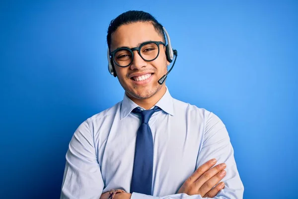 Young brazilian call center agent man wearing glasses and tie working using headset happy face smiling with crossed arms looking at the camera. Positive person.