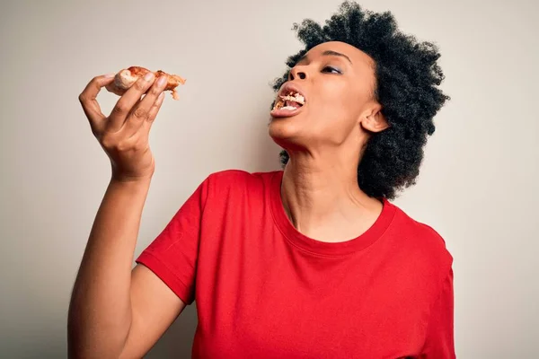 若い美しいアフリカ系アメリカ人のアフロ女性が笑顔で自信を持っています 白い背景の上にイタリアのピザのおいしいスライスを食べる顔に笑顔で立って — ストック写真