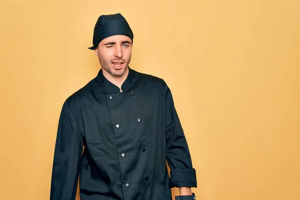 Young handsome cooker man with blue eyes wearing uniform and hat over yellow background winking looking at the camera with sexy expression, cheerful and happy face.