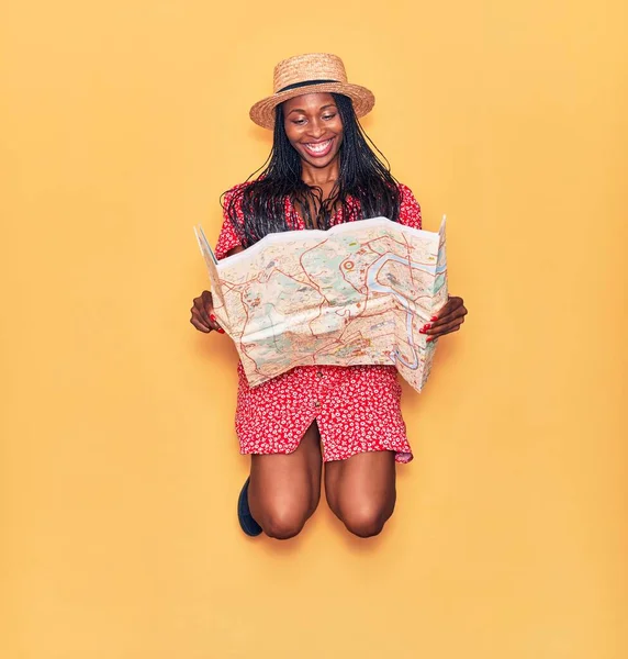 Young beautiful african american woman wearing summer clothes and hat smiling happy. Jumping with smile on face holding city map over isolated yellow background.