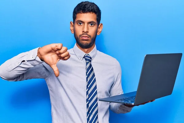 Young latin man holding laptop with angry face, negative sign showing dislike with thumbs down, rejection concept