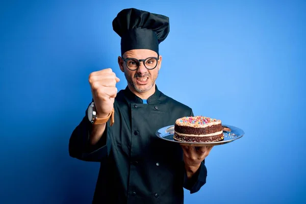 Young handsome baker man wearing cooker uniform and hat holding tray with cake annoyed and frustrated shouting with anger, crazy and yelling with raised hand, anger concept