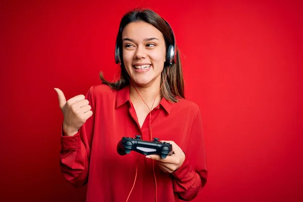 年轻美丽的黑发女孩在玩电子游戏 她用操纵杆和耳机指指点点 带着快乐的面带微笑站在旁边 — 图库照片