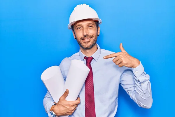 Jovem Bonito Homem Vestindo Arquiteto Hardhat Segurando Plantas Apontando Dedo — Fotografia de Stock