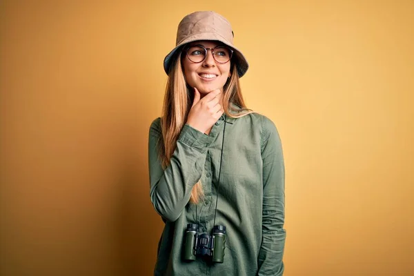 Beautiful blonde explorer woman with blue eyes wearing hat and glasses using binoculars with hand on chin thinking about question, pensive expression. Smiling with thoughtful face. Doubt concept.