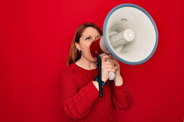 Young beautiful redhead speaker woman screaming communicate using megaphone standing over isolated red background
