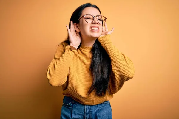 若いブルネットの女性は黄色の隔離された背景の上に眼鏡とカジュアルなセーターを着て耳のジェスチャーで両方の手を聞こうとして ゴシップを探しています 聴覚障害 聴覚障害 — ストック写真