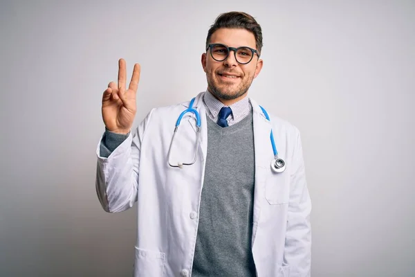 青い目の若い医者の男は 勝利サインをして指を示すカメラを見て笑って孤立した背景の上に医療コートと聴診器を身に着けています 第二番 — ストック写真