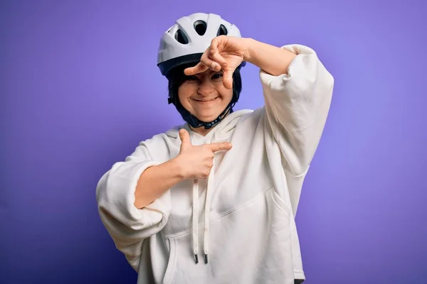 Young down syndrome cyclist woman wearing security bike helmet over purple background smiling making frame with hands and fingers with happy face. Creativity and photography concept.