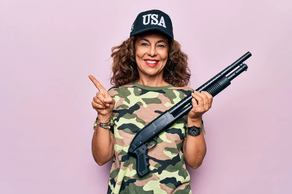 Middle age beautiful hunter woman wearing camouflage t-shirt and usa cap holding shotgun smiling happy pointing with hand and finger to the side