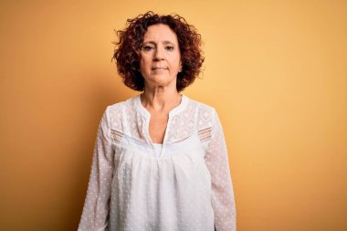 Orta yaşlı güzel kıvırcık saçlı kadın sarı arka planda günlük yazlık elbise giyiyor ve yüzünde ciddi bir ifade var. Basit ve doğal bir şekilde kameraya bakıyorsun..