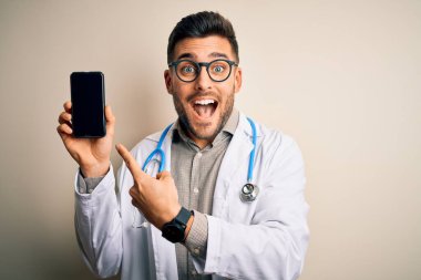 Steteskop takan genç doktor, izole edilmiş arka planda akıllı telefon ekranı gösteriyor. Elle ve parmakla göstererek çok mutlu.