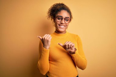 Sarı arka planda süveter ve gözlük takan güzel Afro-Amerikan kızı. Arka tarafı işaret ediyor. El ve baş parmakları havada.