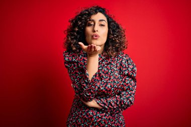 Genç, güzel, kıvırcık, çiçekli bir elbise giyen, kırmızı arka planda dikilmiş kameraya bakıp havada öpücükler saçan, güzel ve seksi bir kadın. Aşk ifadesi.