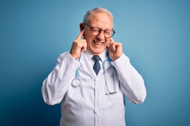 Kıdemli gri saçlı doktor steteskop ve mavi arka planda tıbbi ceket giyiyor. Yüksek sesli müzik sesi için parmaklarıyla kulaklarını kapatıyor. Sağır kavramı.