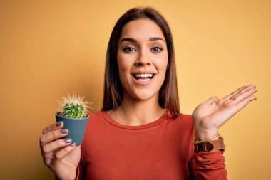 Genç, güzel esmer kadın sarı arka planda küçük bir kaktüs tenceresi tutuyor. Çok mutlu ve heyecanlı. Kazanan ifade zaferi kutluyor.