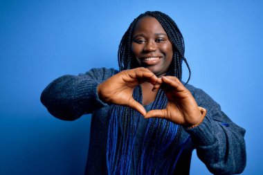 Mavi arka planda normal süveter giyen, elleri ile kalp sembolü şeklinde gülümseyen Afro-Amerikan kadın. Romantik konsept.