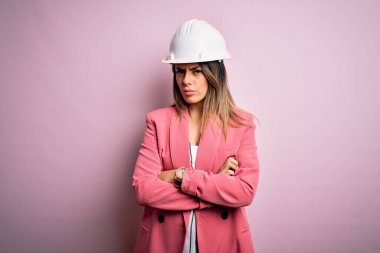 Genç, güzel esmer mimar kadın pembe arka planda güvenlik kaskı takıyor şüpheci ve gergin, yüzünde çapraz kollu tasvip etmeyen bir ifade. Negatif kişi.