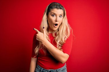 İzole edilmiş kırmızı arkaplanın üzerinde dikilen gündelik tişört giyen genç, güzel sarışın kadın parmağını yana çevirmiş, ağzı açık şaşkın bir ifade sergiliyor..