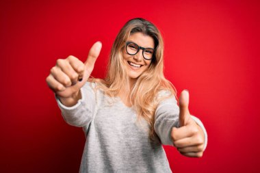 Güzel, sarışın, süveter ve gözlük takan genç bir kadın, izole edilmiş kırmızı arka plan üzerinde olumlu bir jest yapıyor, elleriyle gülümsüyor ve başarı için seviniyor. Kazanan jest.