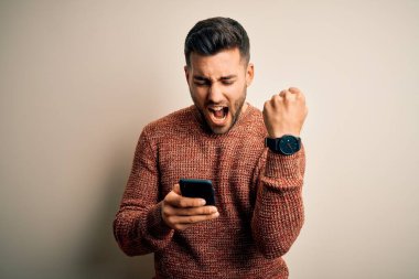 Beyaz arka plan üzerinden akıllı telefon kullanan genç bir adam sinirlenmiş ve sinirlenmiş bir şekilde bağırıyor, çıldırmış ve el kaldırarak bağırıyor.