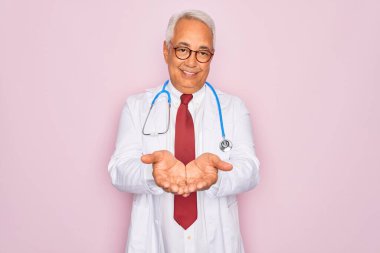 Orta yaşlı, kır saçlı, steteskop takan ve elleri avuçlarında gülümseyen, el hareketi yapan uzman doktor. Tutun ve koruyun
