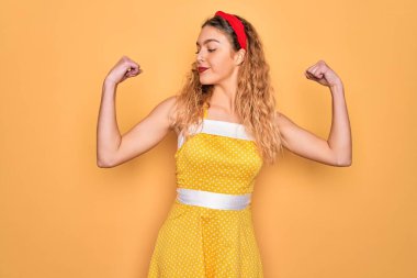 Sarı arka planda dikilen mavi gözlü güzel sarışın kadın gururla gülümseyen kol kaslarını gösteriyor. Fitness konsepti.