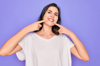 Genç, güzel, esmer bir kadın mor arka plan üzerine günlük beyaz tişörtler giyip neşeli bir şekilde gülümseyerek parmaklarını ve ağzını işaret ediyor. Diş sağlığı konsepti.