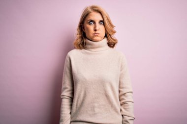 Orta yaşlı, güzel, sarışın bir kadın pembe arka planda komik suratlı, gelişigüzel balıkçı yaka kazak giyiyor. Havayla şişmiş ağız, çılgın bir ifade..