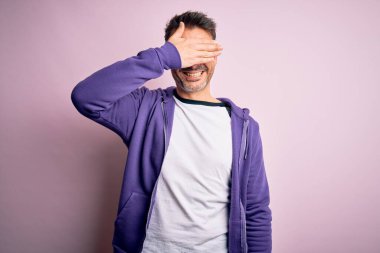 Mor eşofman altı giyen ve pembe arka planda duran gözlüklü yakışıklı bir adam. Gülümsüyor ve yüzünde şaşkınlık için gözleri kapatan bir el ile gülüyor. Kör kavram.