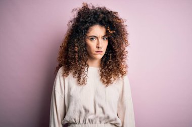 Kıvırcık saçlı ve piercingli güzel bir kadın pembe arka planda sıradan bir süveter giyiyor şüpheci ve gergin, sorun yüzünden suratını asıyor. Negatif kişi.