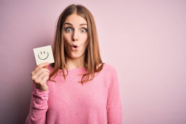 Genç, güzel, kızıl saçlı bir kadın elinde gülümseme kağıdıyla şok olmuş, korkmuş ve korku ifadesinden heyecanlanmış emoji mesajı tutuyor.