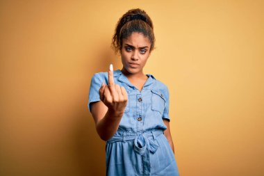 Young beautiful african american girl wearing denim dress standing over yellow background Showing middle finger, impolite and rude fuck off expression