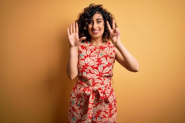 Genç, güzel, kıvırcık Arap kadın tatilde. Çiçekli bir elbise ve güneş gözlüğü takıyor. Yedi numaralı parmaklarıyla işaret ediyor ve gülümsüyor..