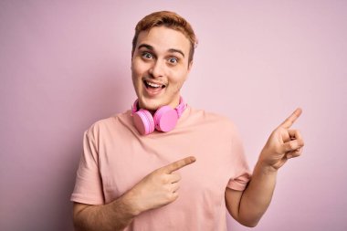 Genç yakışıklı kızıl adam müzik dinliyor pembe arka planda kulaklık kullanıyor gülümsüyor ve kameraya bakıyor iki eliyle ve parmaklarıyla kenarı işaret ediyor..