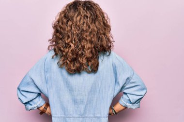 Orta yaşlı güzel bir kadın pembe arka planda dikilmiş sıradan bir kot gömlek giyiyor geriye doğru bakıyor kolları vücudunda.